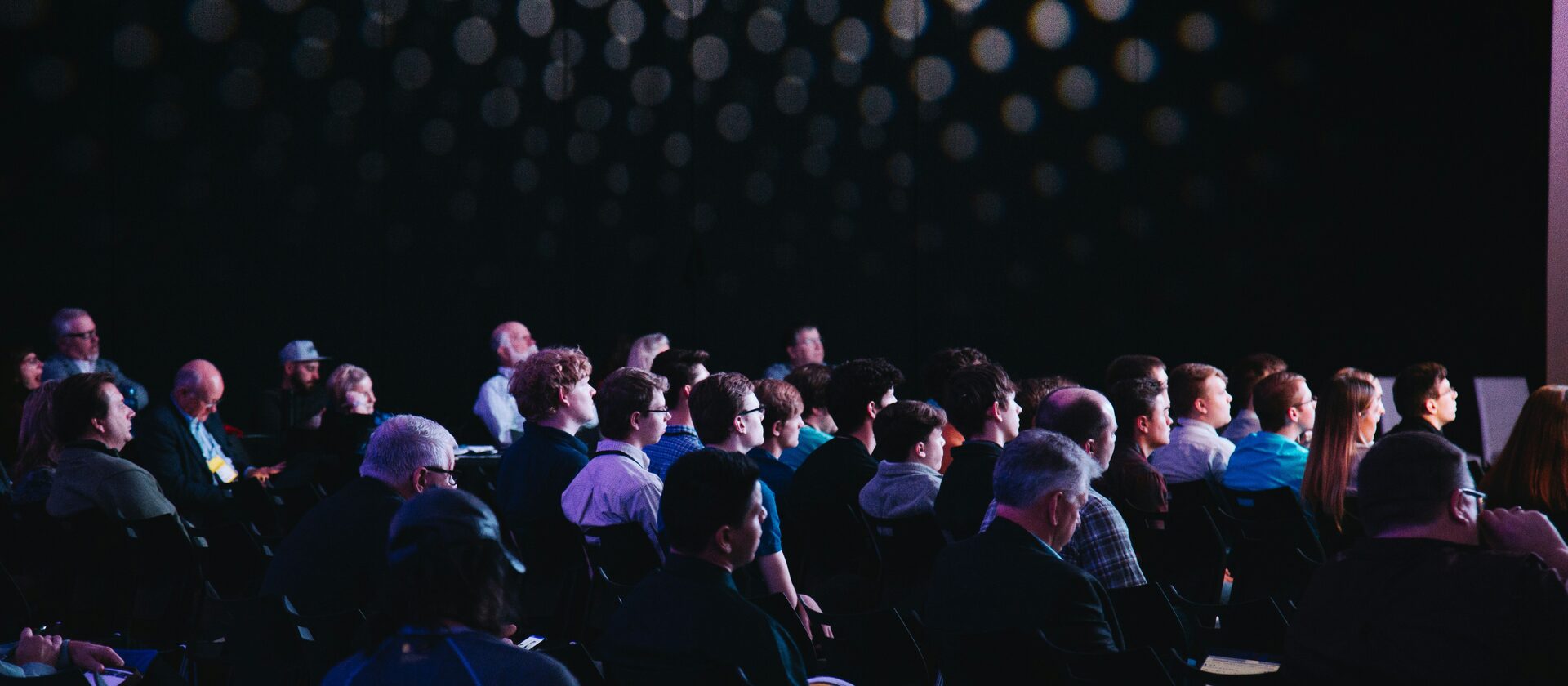 An audience seated in a darkened auditorium, lights above the stage